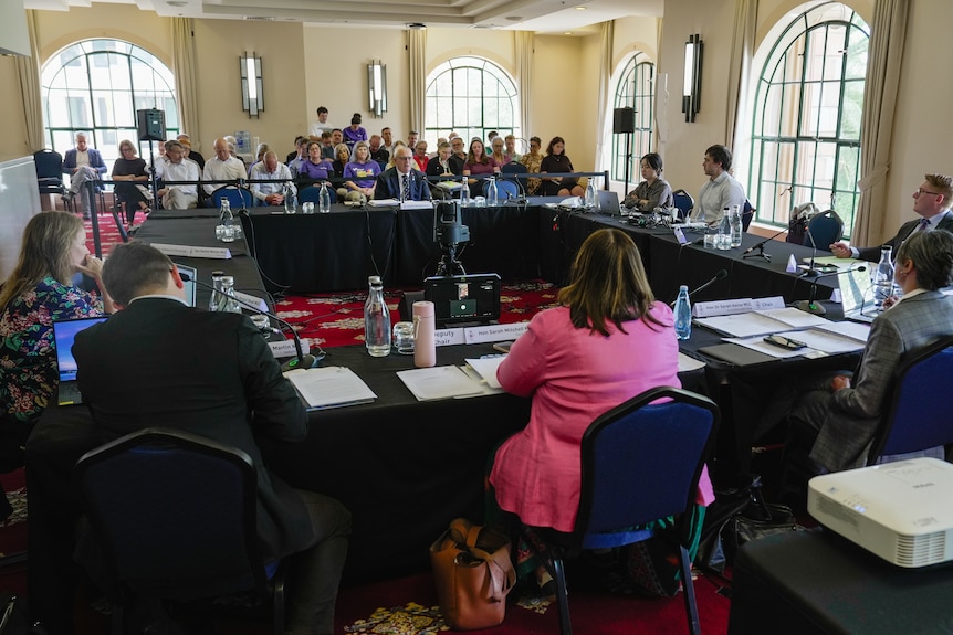 Tables set up in a square formation for an inquiry hearing with an audience in the background.
