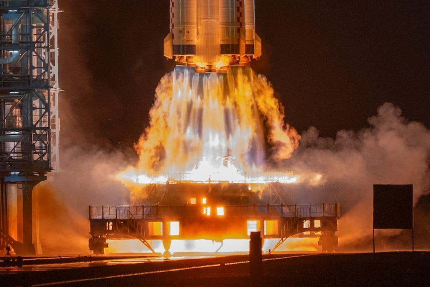 Fiery spaceship boost thrusters illuminated in light blue and orange flames above a space station launchpad.