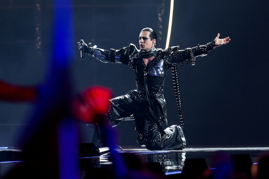 A performer dressed in gothic balck kneels on stage holding out their arms