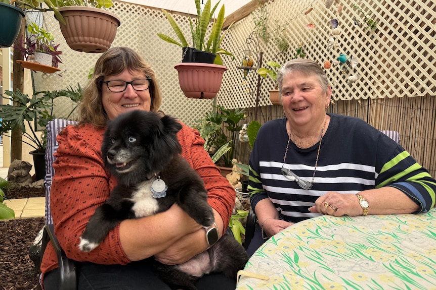 A bespectacled brunette woman, Rhonda, holds a fluffy black and white dog, next to grey-haired woman Carol.