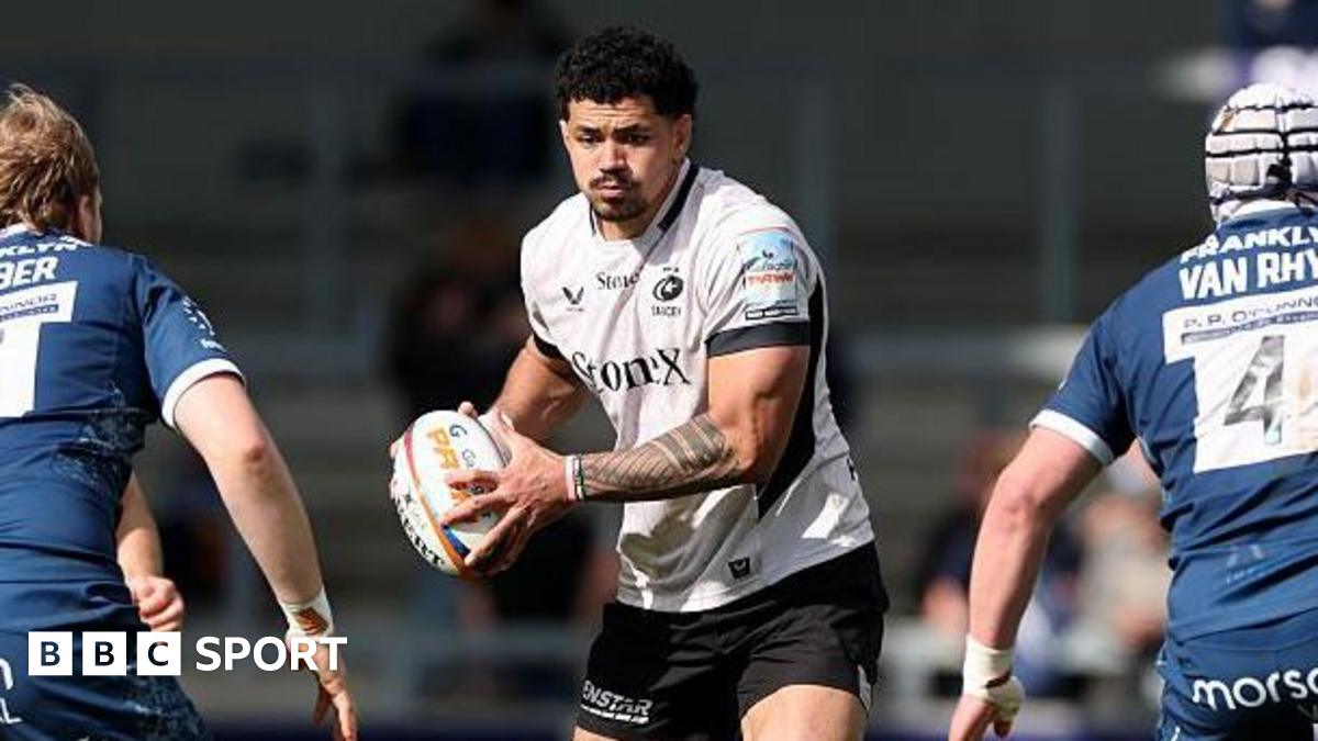 Theo McFarland (centre) stands between two Sale players holding a rugby ball in two hands during a game for Saracens