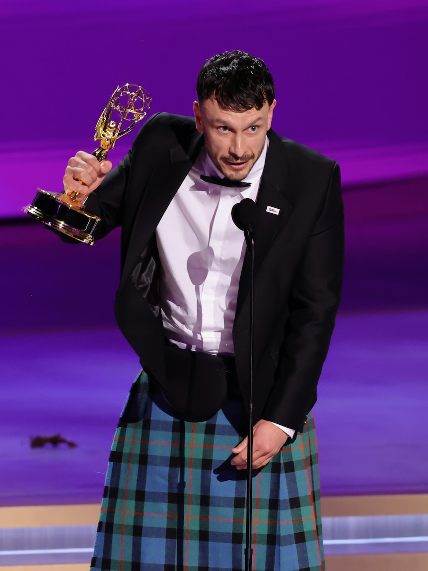 A man wearing a tuxedo with a kilt holds an Emmy trophy while standing onstage and speaking into a microphone.