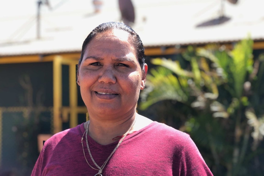 Patricia Riley, an Indigenous woman, stands in front of her house, which has satellite dishes. She wears a burgundy t-shirt.