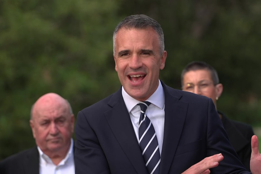 Premier Pere Malinauskas is wearing a suit and tie as he addresses media in an area flanked by green trees.