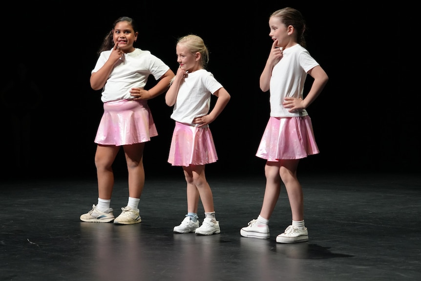 Three girls wearing white shirts and sparkly pink skirts pose with their hands on their chin on a black stage.