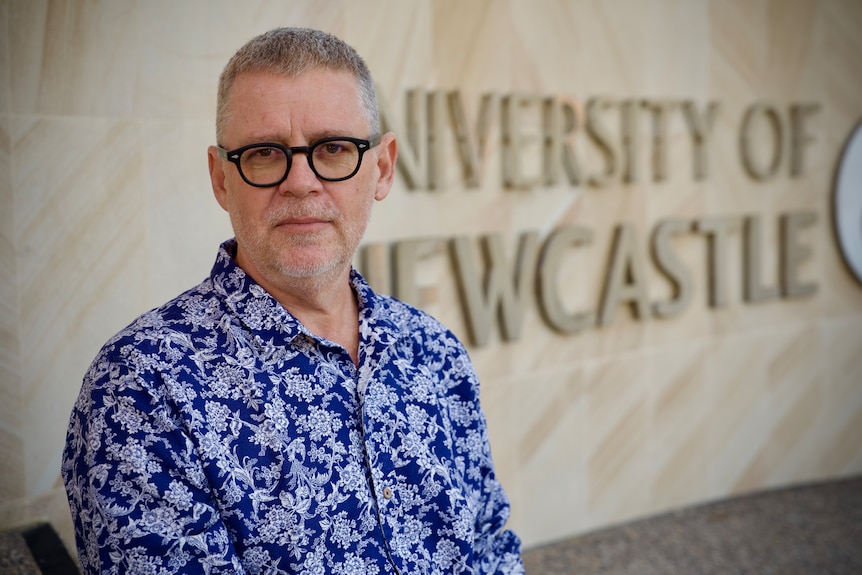 A man in a blue and white patterned shirt sits in front of a University of Newcastle sign.