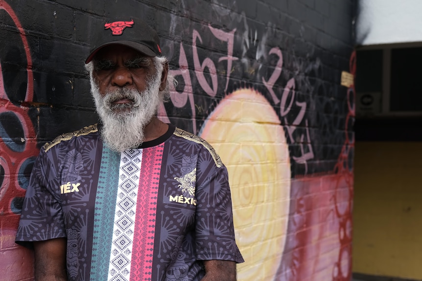 Paul Andy, standing in front of an Aboriginal flag mural, looking at the camera.