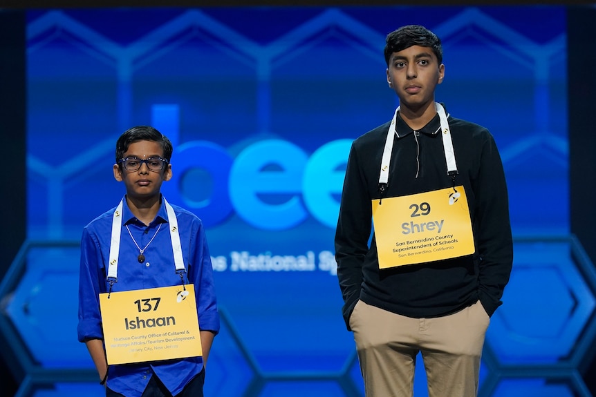 Two adolescent boys stand on a stage with signs displaying their names around their necks