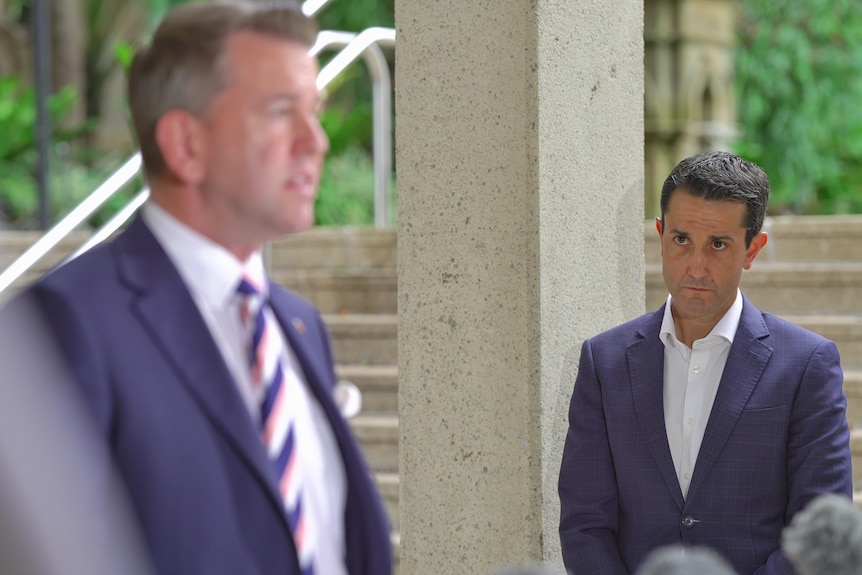 A man in a blue suit leaning on a pole watching another man speak.