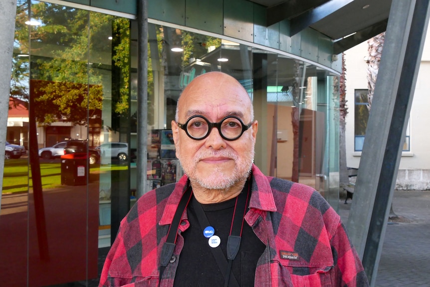 An older man with tan skin and wearing glasses wearing a red and black flannel shirt standing in front of a glass building.