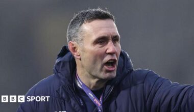 Stephen Jones pointing with his left hand towards players out of shot, while wearing a lanyard and a thick padded coat