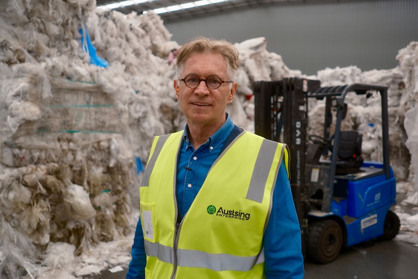 A white haired man with glasses, a blue collared shirt and yellow vest stands near a blue forklift and piles of plastic.