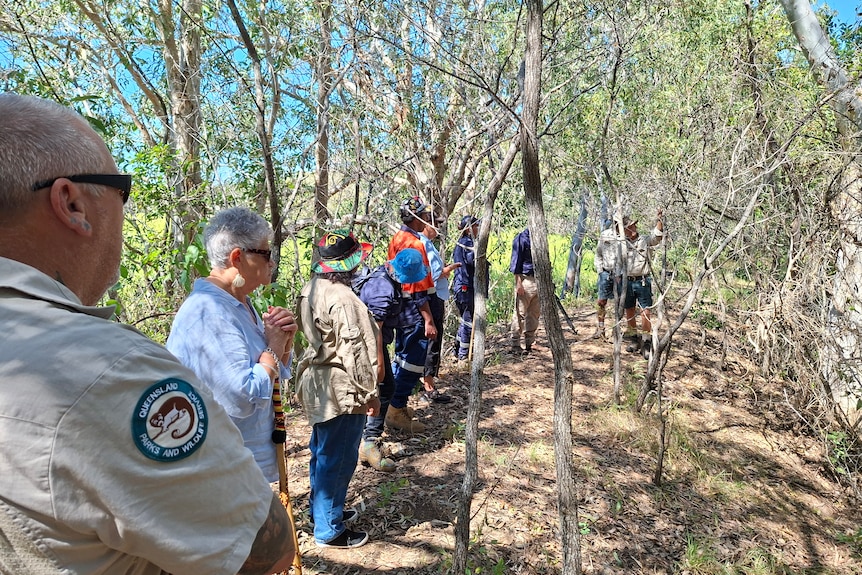 A group of people stand along along a mound of dirt, surrounded by trees.