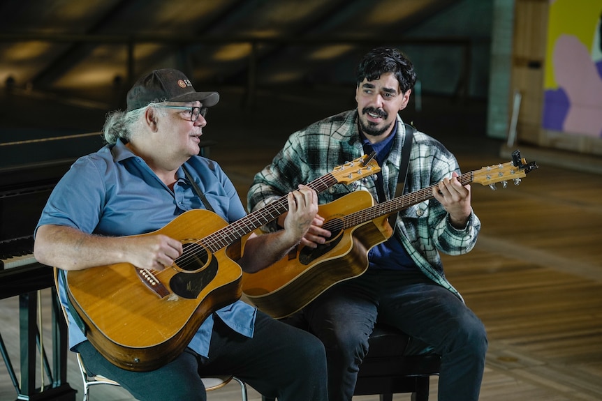 Goori and Buddy sit side by side in a room with wooden floors, playing guitars.
