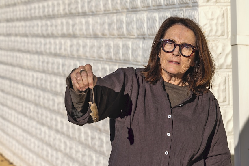 Karen is wearing a brown shirt and holds a mouse by its tail with the street behind her.