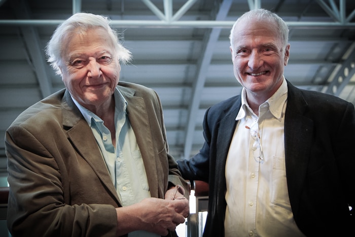 Two older man with white hair in jackets standing side by side shaking hands.