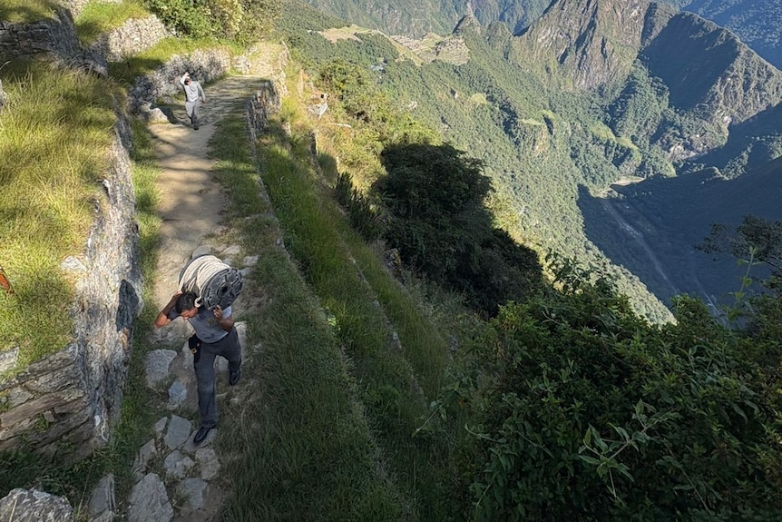 A Peruvian man in grey clothing carrying equipment on his shoulders on a stony path in the jungle.