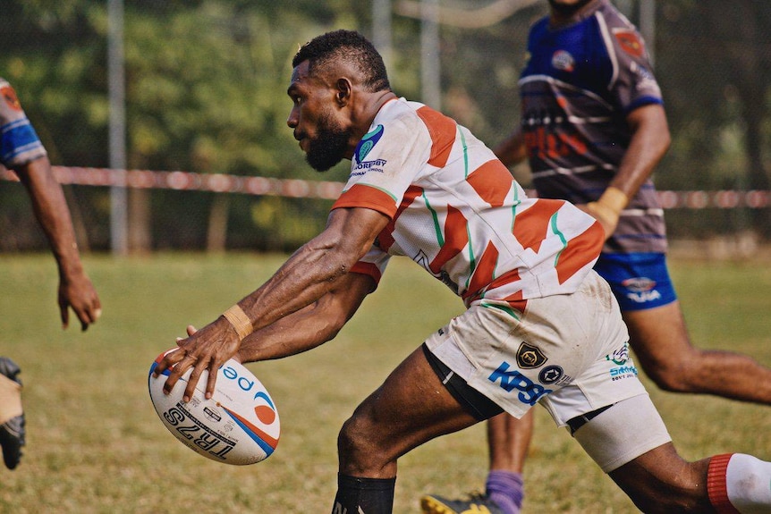 A Papua New Guinea man holding a rugby ball running on a field wearing white and red stripe rugby jersey and white shorts