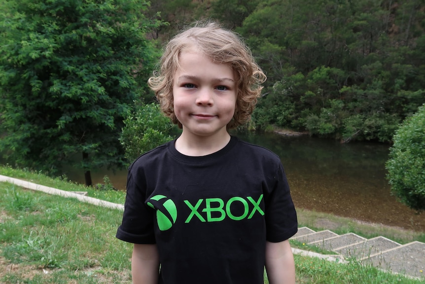 A young boy smiles at the camera wearing a black shirt with the words XBox on it.