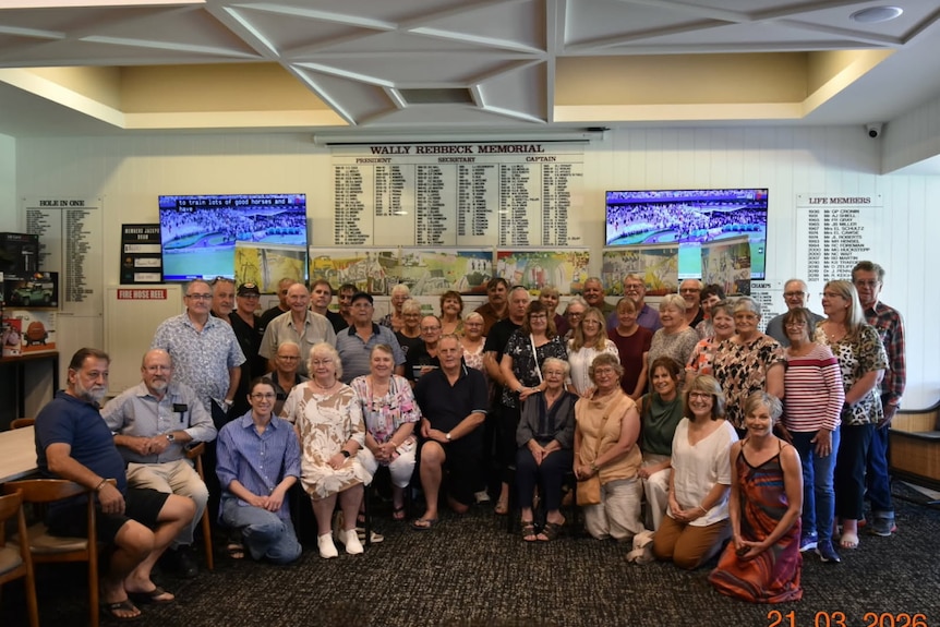 About 70 people stand and sit smiling in front of a banner at a golf club.