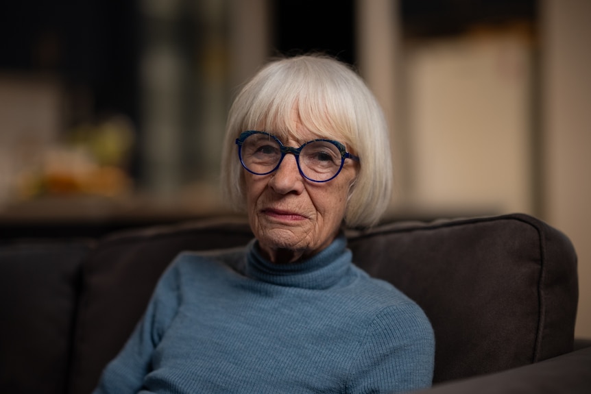 An older woman with a white-grey bob and fringe with large blue-framed glasses and a blue turtleneck sits on a couch.