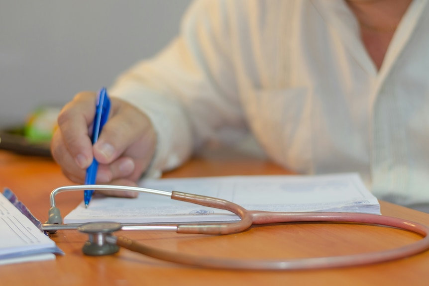 A female doctor writing in a note pad, with a stethoscope on the table.