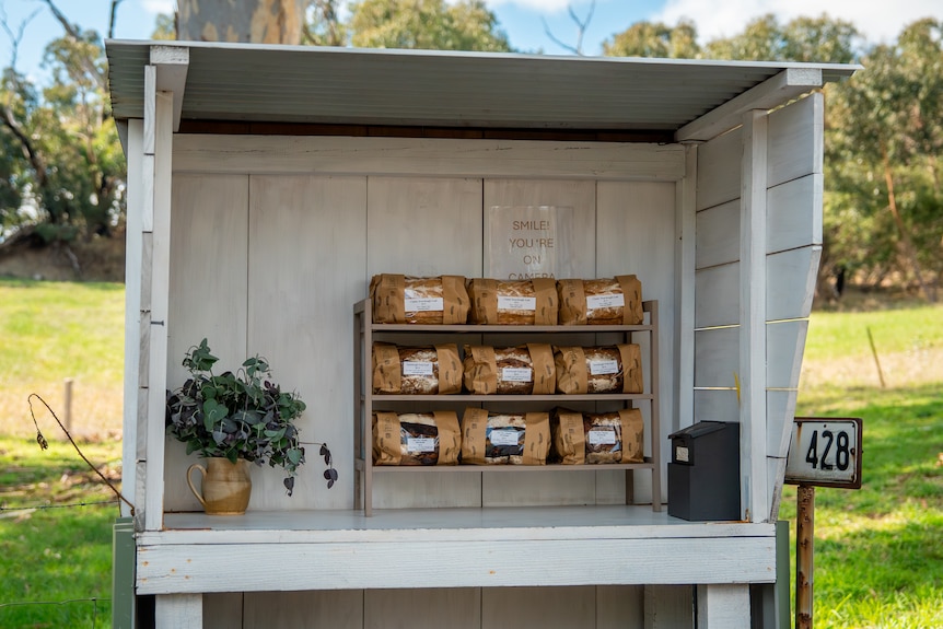 A white wooden stall stands in a grassy area, it is stocked with loaves of bread in paper packaging