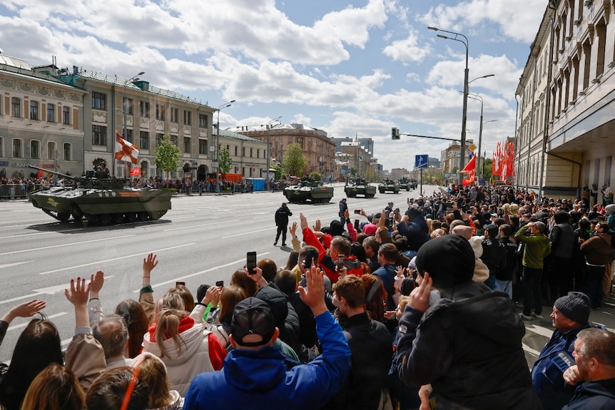 Tanks driving down a road, as a large crowd cheers.