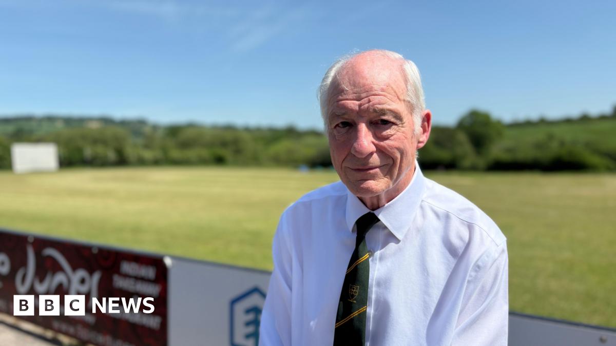 Tony Evans, a man with white hair, sits with his back to the cricket match, wearing a white shirt and green and yellow Purnell Cricket Club tie.