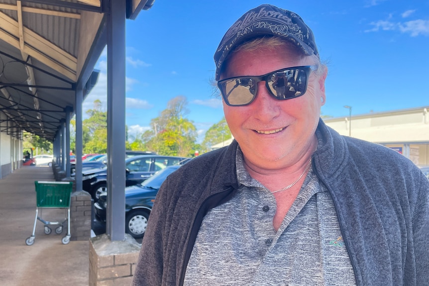 A man wearing a cap and sunglasses stands outside a supermarket with a smile on his face