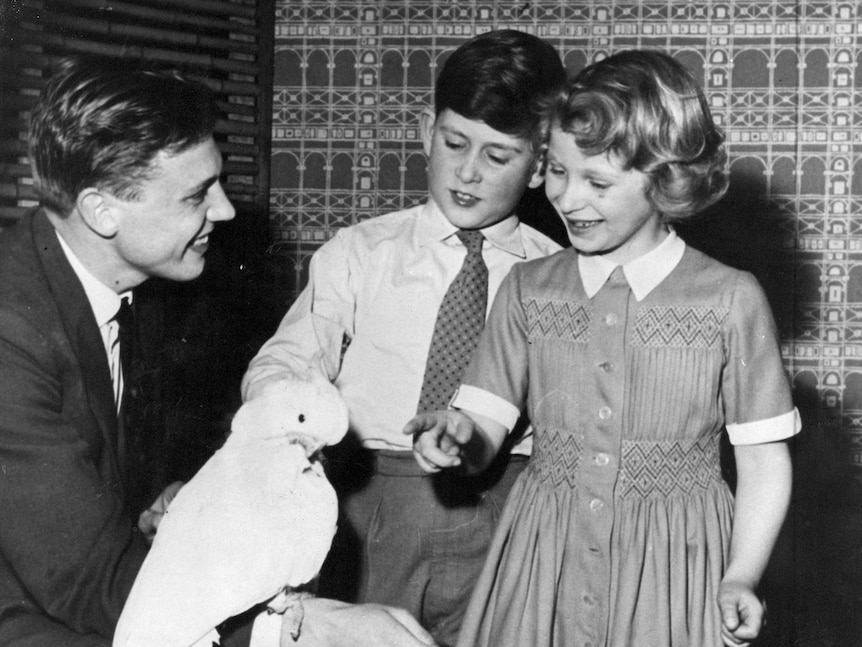 Black and white photo of David Attenborough holding a cockatoo in front of a small boy and girl.