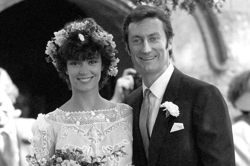 Black and white image showing bride and groom standing, smiling. Woman in a bridal dress and flower crown holds a bouquet