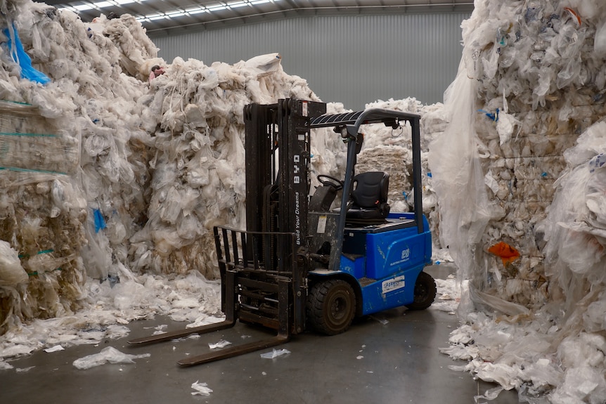 A blue and black forklift is parked inside a shed surrounded by piles of clear and white soft plastic.