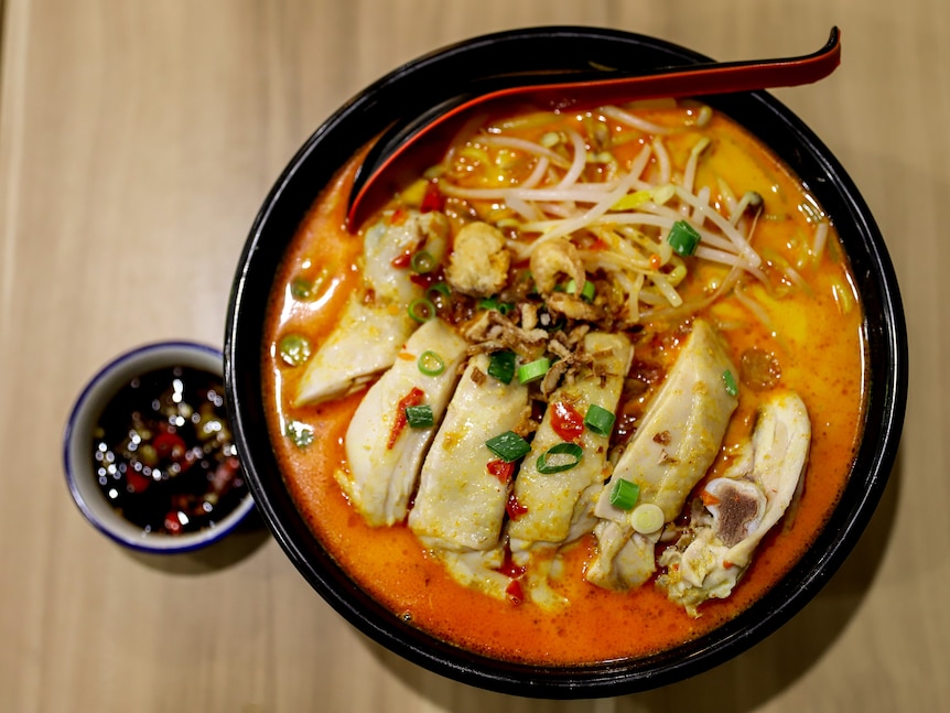 A large bowl containing with orange liquid, a spoon, chicken meat, bean sprouts, pieces of chilli and spring onion sits on a tab