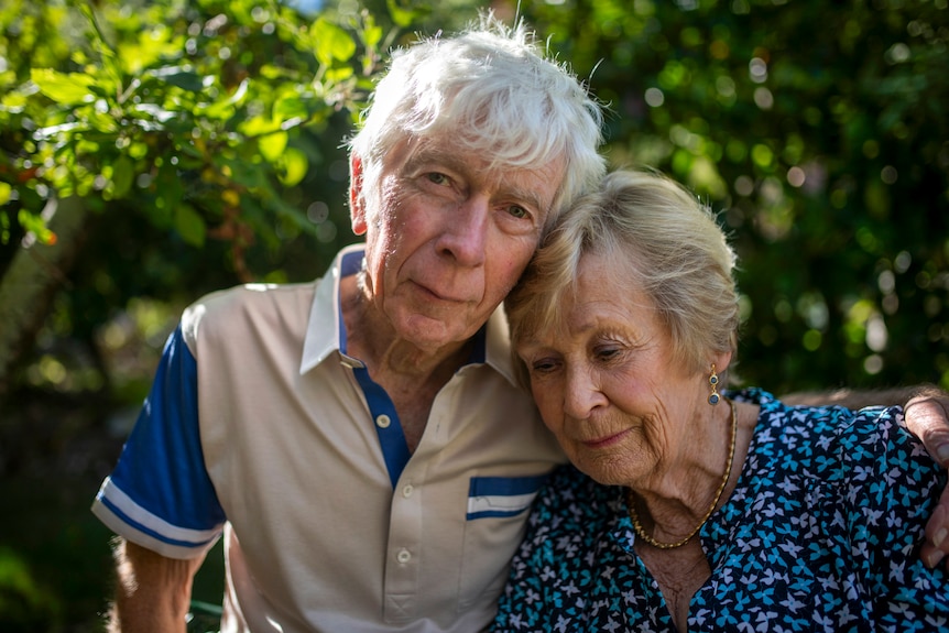 An elderly gentleman looks into the camera with his arm around his wife in dappled light in the green garden.