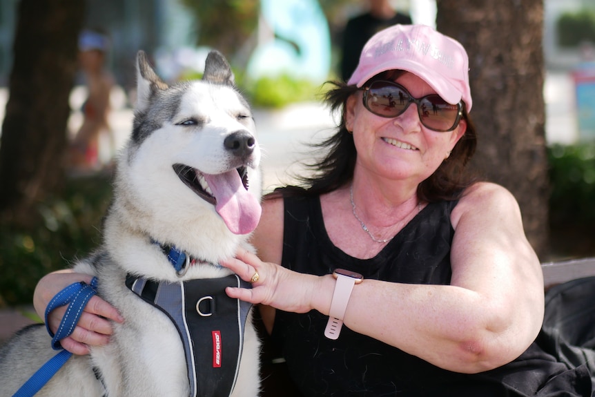 A woman wearing a pink cap with a husky dog sits outside on a park bench and smiles at the camera.