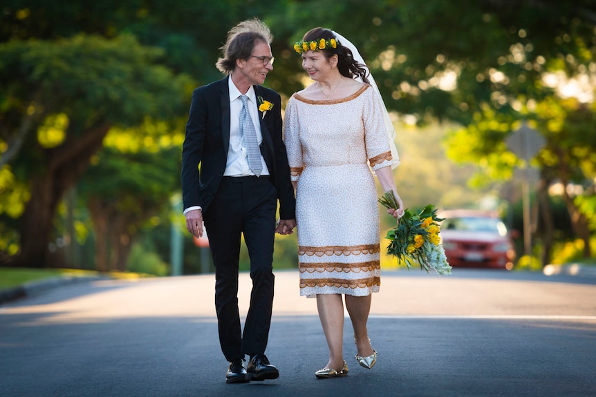 Mick Medew wears a black suit and white short and Ursula Collie wears a white dress on their wedding day. Both are smiling.