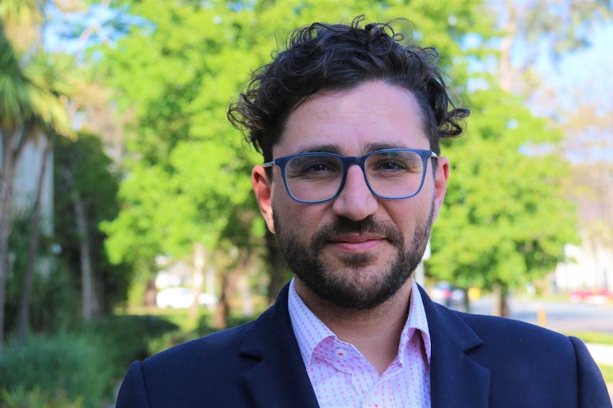 A bespectacled, bearded man in a dark suit with no tie looks into the lens.