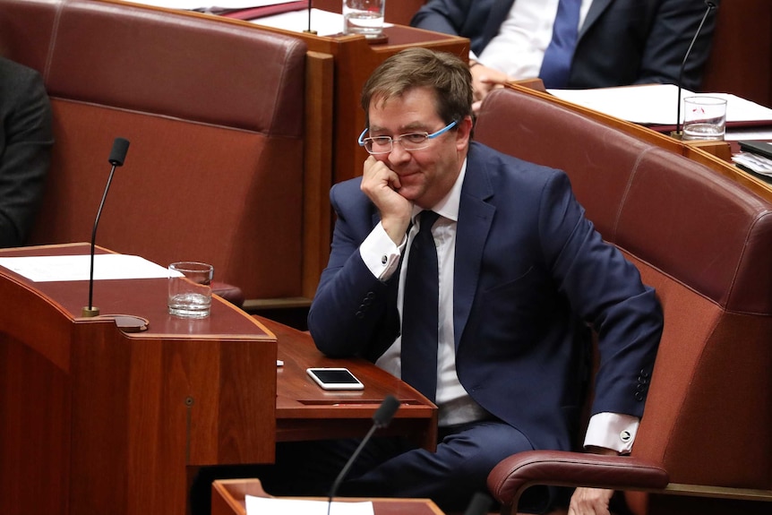McGrath is wearing a blue suit, hand under chin, smiling.