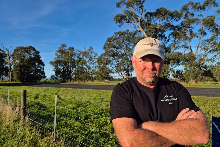 A middle aged man in a black tshirt and beige cap stands with crossed arms in a field, in front of a wire fence.