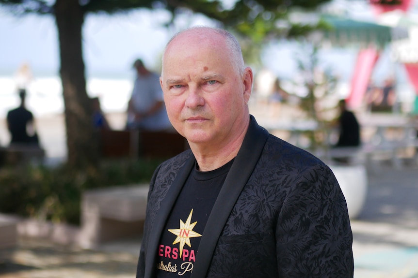 A man wearing a black jacket stands outdoors with a serious expression on his face.