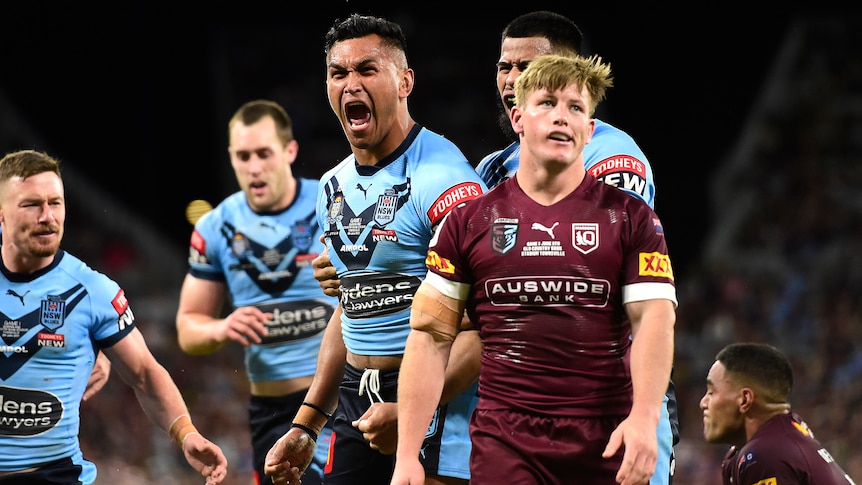 A NSW State of Origin player roars in celebration as a Queensland player looks dejected after a try.