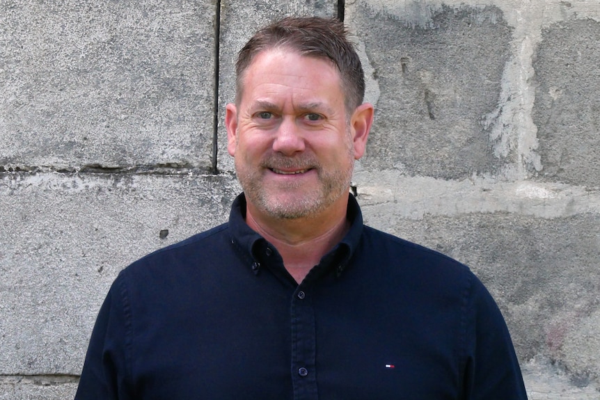 A man with brown-grey hair and stubble wearing a dark blue collared shirt stands in front of a grey wall.