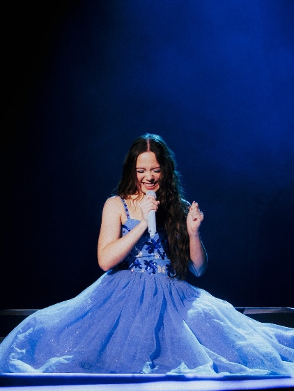 Laufey in a big blue dress, smiling and performing on stage.