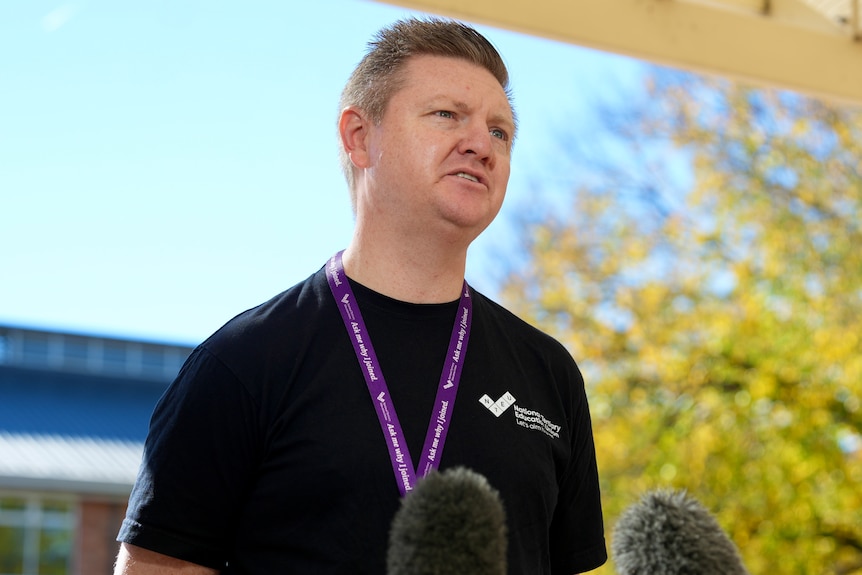 A man with short hair stands outdoors looking serious.