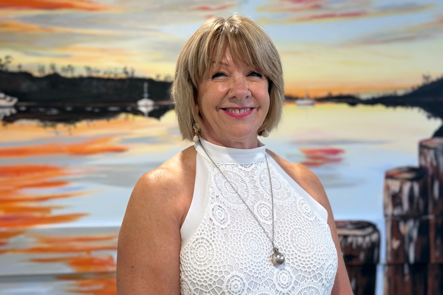 A woman in a white singlet with a blonde bob smiles as she stands infront of a colourful painting.