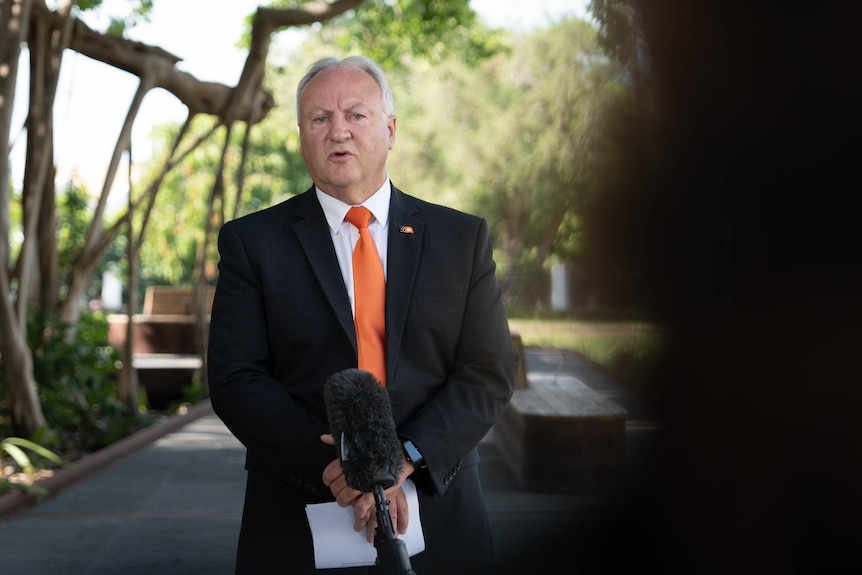 A man in a suit an orange tie speaks to the media