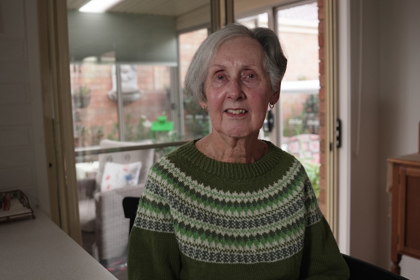 A woman with grey hair and green jumper sits at her kitchen bench.