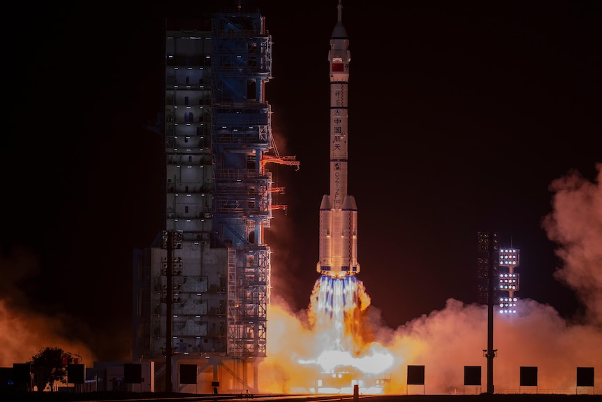 A white space ship launching alongside a vertical control tower, above a bright orange ball of flame and smoke cloud.