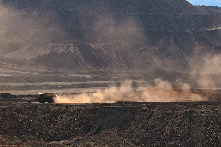 A mining truck drives in the distance a large cloud of dust behind it. It is dwarfed by a large mountain in a mine site.